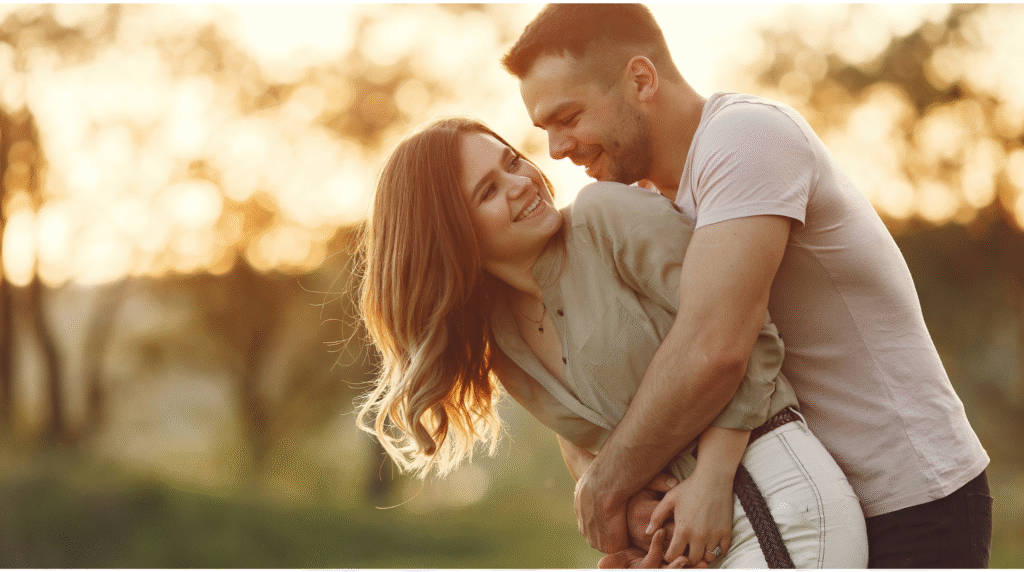 Casal jovem sorrindo e se abraçando carinhosamente em um parque ao pôr do sol, representando o cuidado e a conexão no relacionamento.