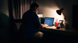 Homem de óculos e camiseta azul sentado em uma mesa, digitando em um laptop em um quarto escuro, com uma luminária acesa ao lado. A cena sugere isolamento e uso prolongado de tecnologia.